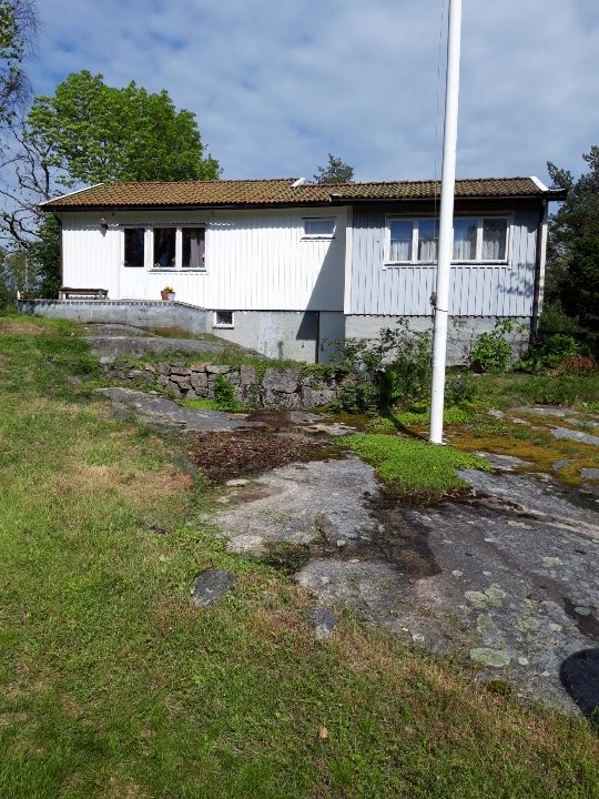 The exterior of the cottage is shown, featuring white paneling and a sloped roof. A small porch with a bench is visible at the entrance. Surrounding greenery enhances the natural setting, while a large rock formation lies in the foreground. Soft light brightens the scene.