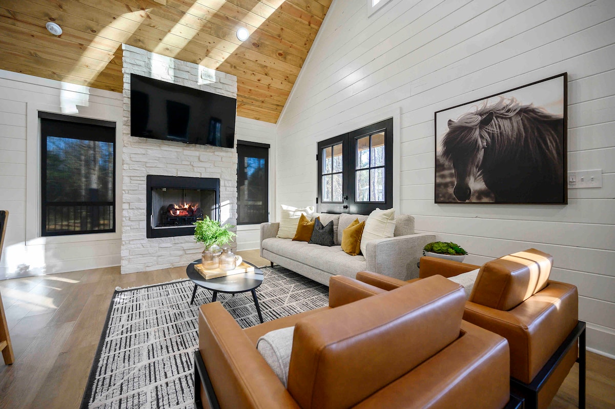 A spacious living area features a white shiplap wall and a stone fireplace. A large television hangs above the fireplace, while natural light brightens the room through dark-framed windows. A cozy couch with decorative cushions and two brown armchairs are placed on a patterned area rug.