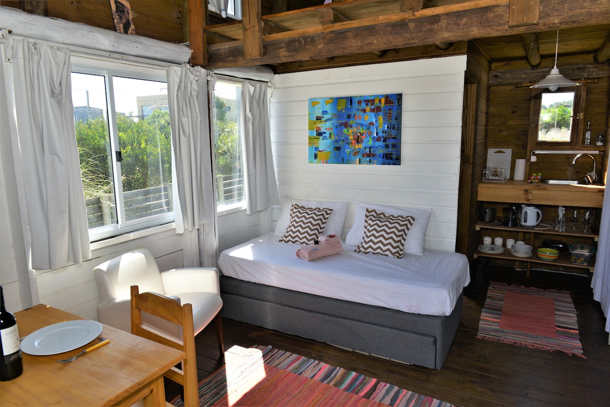 An open-concept living space displays a cozy arrangement with a convertible bed and light-colored decor. A wooden dining table with chairs complements the space. Bright natural light streams in through large windows, highlighting a colorful wall art piece and a kitchenette area featuring various kitchenware.