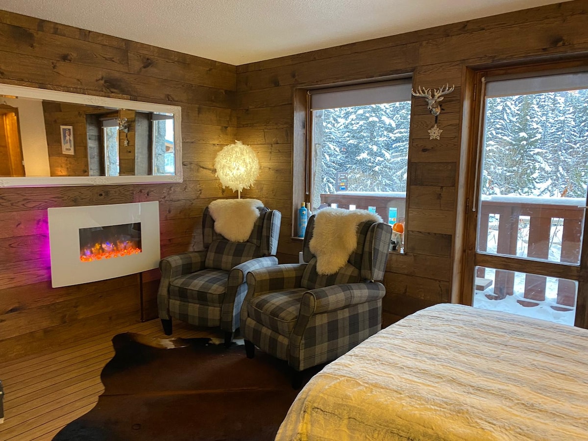 A cozy room features two plaid armchairs positioned near a decorative electric fireplace. Natural light filters through large windows showcasing snowy outdoor scenery. The wooden walls and a soft area rug add warmth to the inviting interior design.