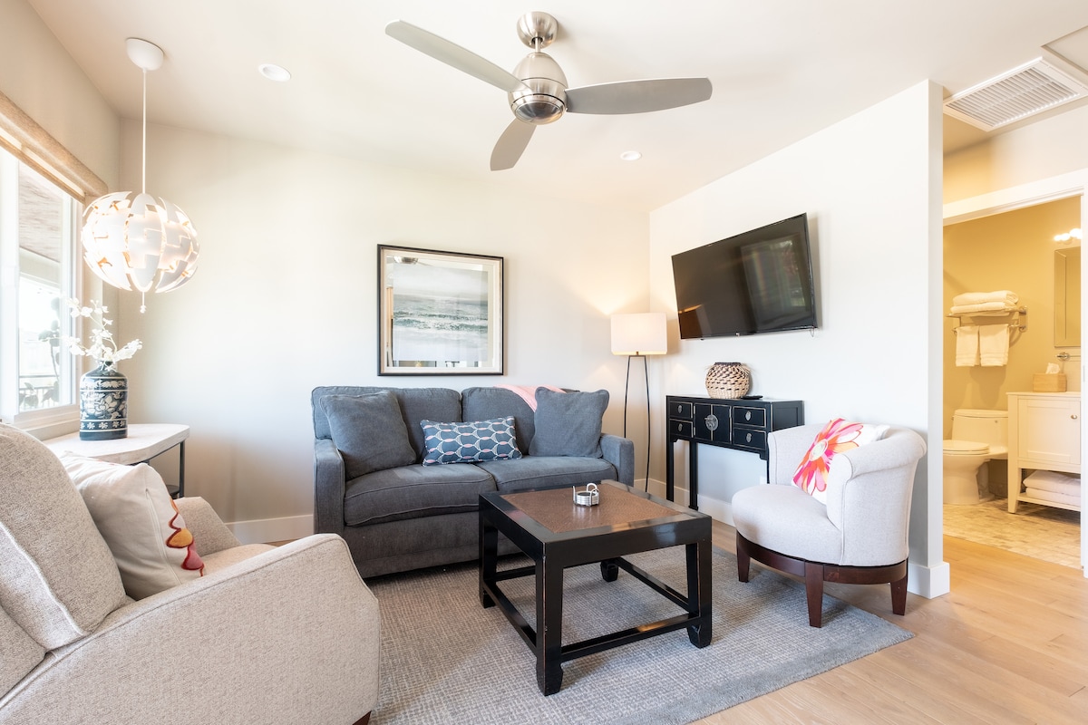 A living room features a comfortable gray sofa, a light-colored armchair, and a coffee table atop a textured area rug. Natural light filters through large windows, and a ceiling fan is visible above. A wall-mounted television is placed opposite the seating area.