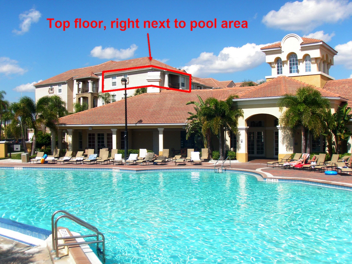 The image depicts a top-floor condo situated adjacent to a large outdoor pool area. The pool features a clean, inviting water surface and lounge chairs arranged along the poolside. The clubhouse, characterized by its Mediterranean-style architecture, is in the background, surrounded by palm trees.