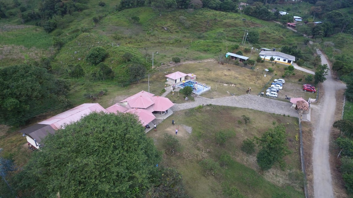 An aerial view captures a rural property featuring several structures with pink roofs, a swimming pool, and ample green space. Nearby, organized parking areas are visible, along with walking paths and natural landscapes showcasing the surrounding hills and lush vegetation.