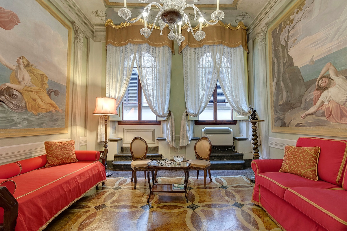 The living room displays original frescoes illustrating serene scenes on either side. Two red sofas frame a small table, complemented by an elegant chandelier overhead. Natural light filters through large windows, draped with delicate lace curtains that add to the room's charm.