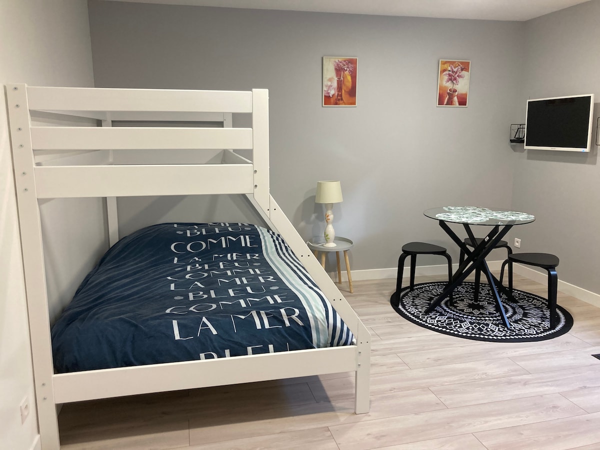 A compact studio featuring a loft bed with a patterned blue duvet cover. A small round dining table with two black chairs is positioned beside the bed. A lamp and a wall-mounted TV are visible, along with framed floral artwork on the gray walls.
