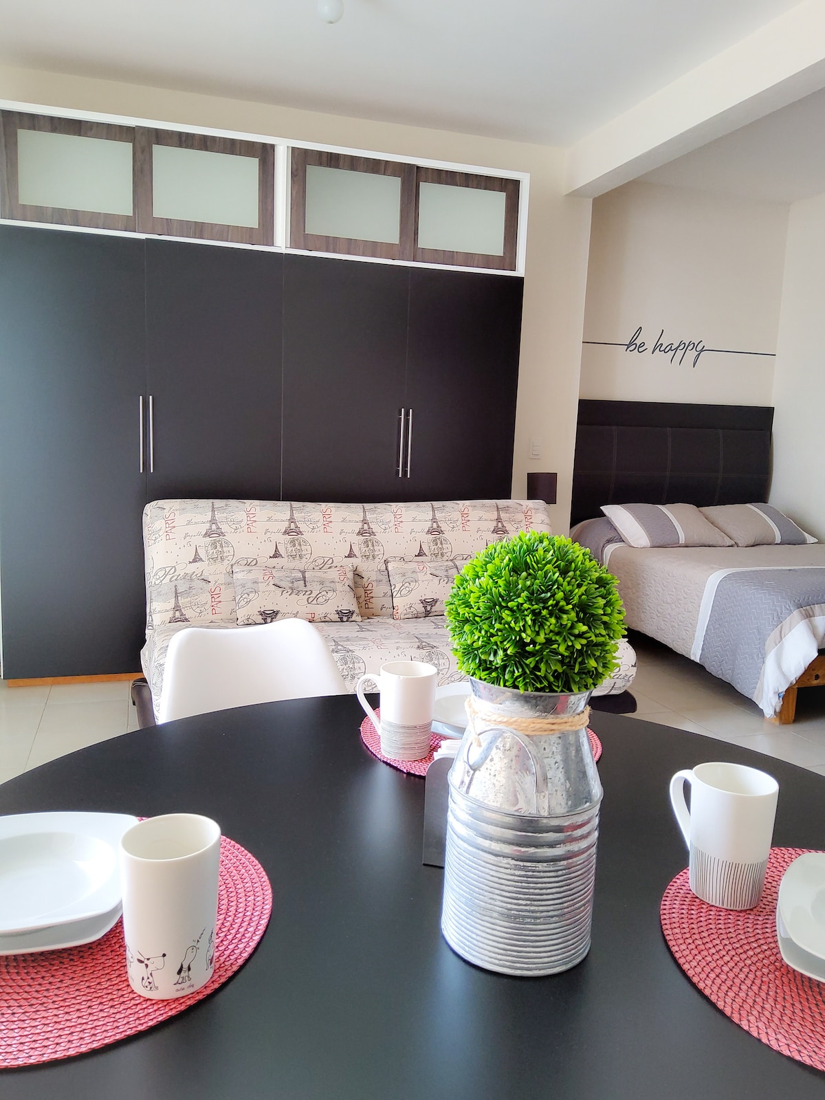 A modern living space is highlighted, featuring a table set with plates and cups. A decorative plant sits in a metal vase at the center. In the background, a sofa with an Eiffel Tower motif and a bed with neutral bedding are visible, complemented by wall art that reads "be happy."