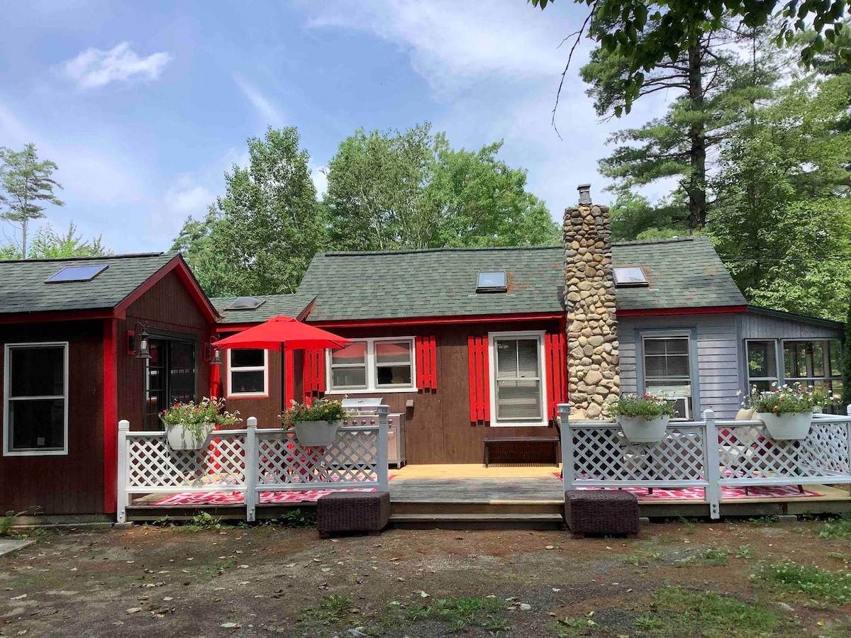 A welcoming cabin exterior features a stone chimney and large windows, allowing natural light to fill the interior. A porch adorned with red umbrellas and flowering planters invites relaxation. The rustic structure is surrounded by lush greenery, emphasizing its tranquil wooded setting.