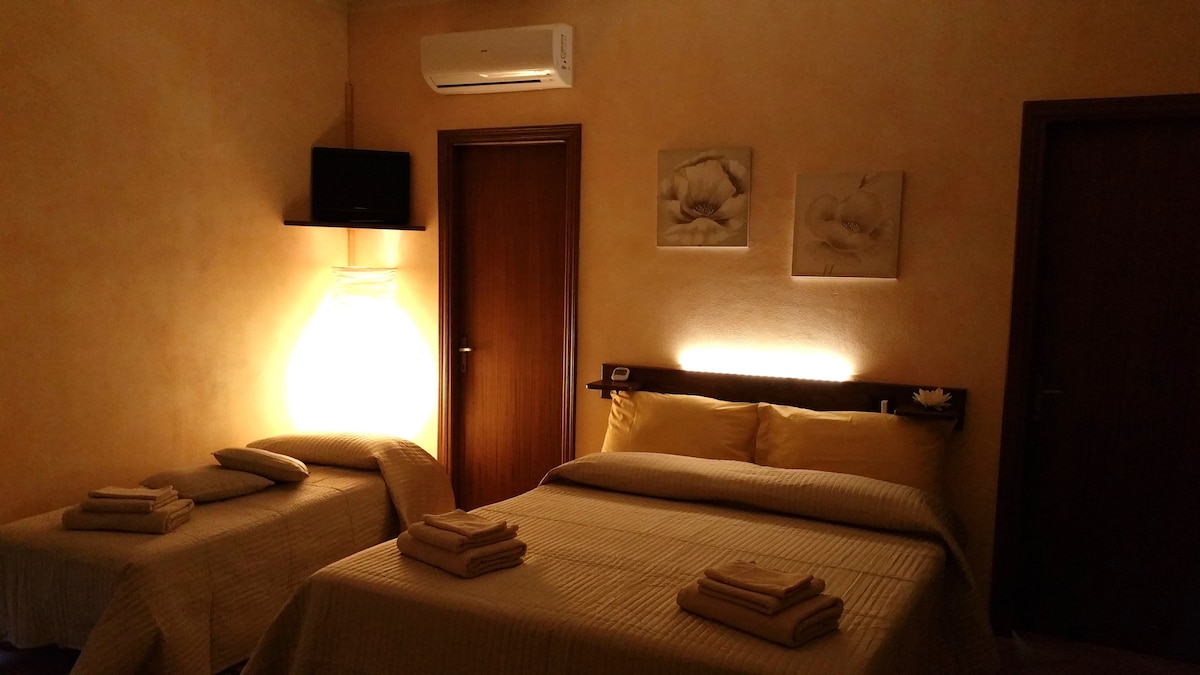 A warm-toned room features a spacious bed and a single bed, both with neatly arranged linen. Soft lighting from a floor lamp and wall-mounted TV creates a cozy ambiance. Floral wall art complements the simplicity of the decor, with wooden doors adding a natural touch.