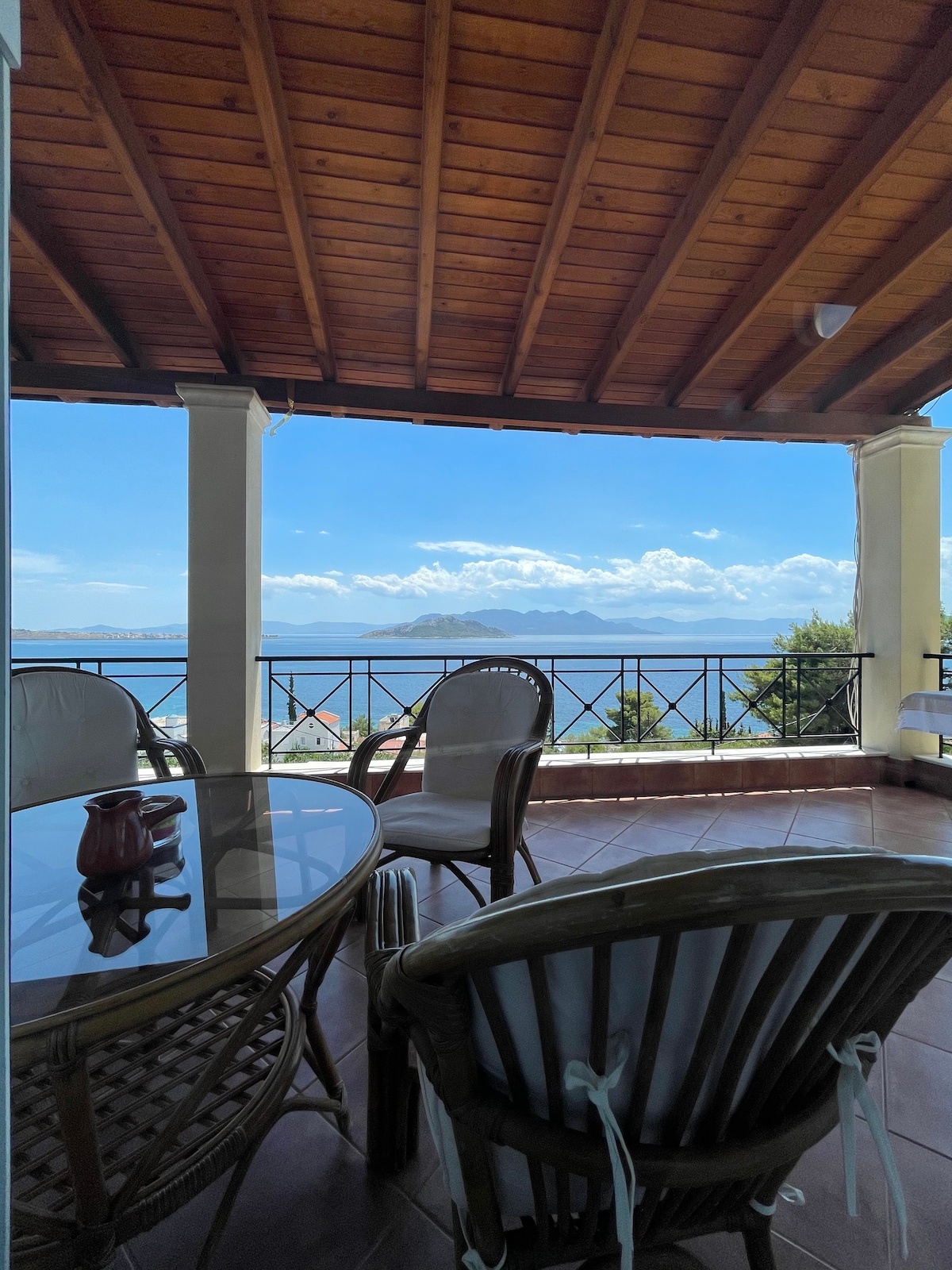 A covered balcony features a round glass table surrounded by wicker chairs. The sea and horizon are visible in the background, framed by the wooden beams of the ceiling and the delicate railing. Natural light filters in, creating a calm outdoor space.