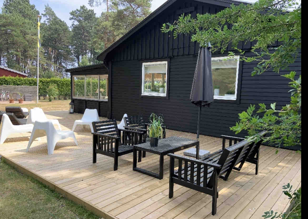 A large wooden deck surrounds the black cabin, featuring outdoor seating arrangements. Black wicker furniture is paired with a small table, and a parasol provides shade. The space is framed by greenery and large windows, contributing to a relaxed outdoor atmosphere.