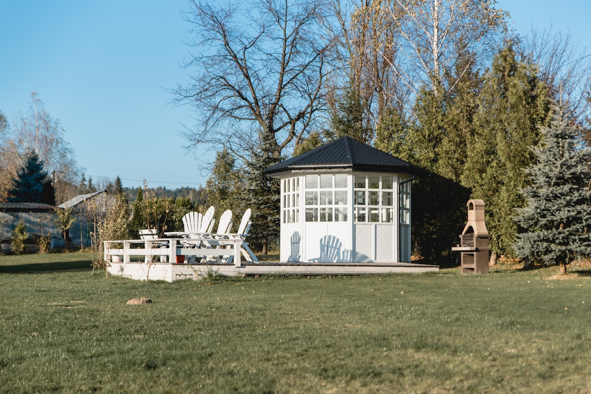 A charming gazebo is situated in a spacious garden, surrounded by greenery. A wooden deck is furnished with light-colored adirondack chairs. Nearby, a traditional barbecue grill stands ready for outdoor cooking, enhancing the inviting atmosphere of the outdoor space.