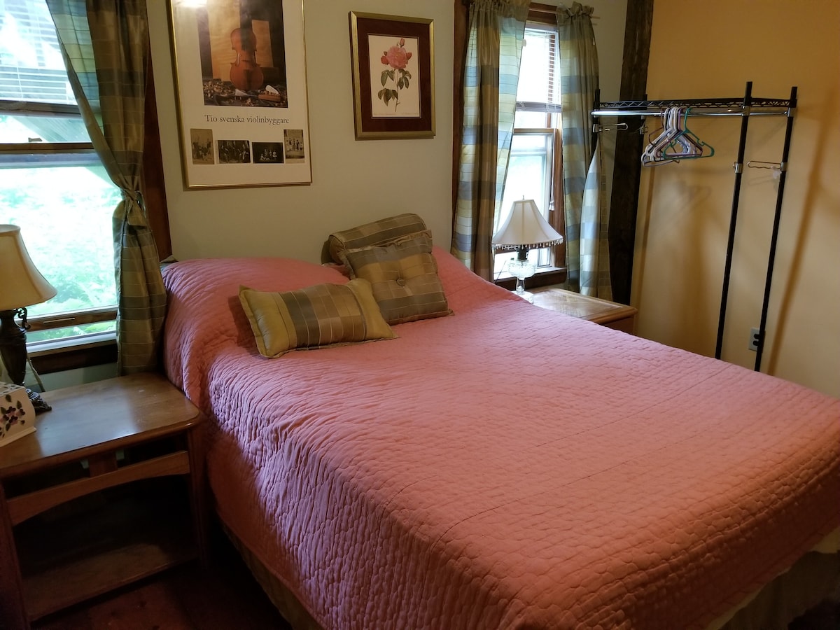 A spacious bed with a light pink quilt is positioned against a wall, surrounded by two windows adorned with patterned curtains. A bedside table is located on either side, with lamps providing soft lighting, and a clothing rack is visible along one side.
