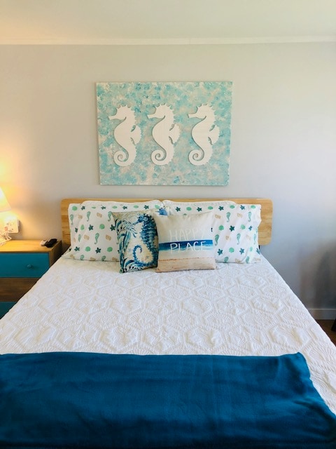 A cozy sleeping area is highlighted by a queen bed adorned with a white quilt and decorative pillows in ocean-inspired patterns. The wall is decorated with a coastal-themed artwork featuring seahorses, and a nightstand with a lamp provides gentle lighting.