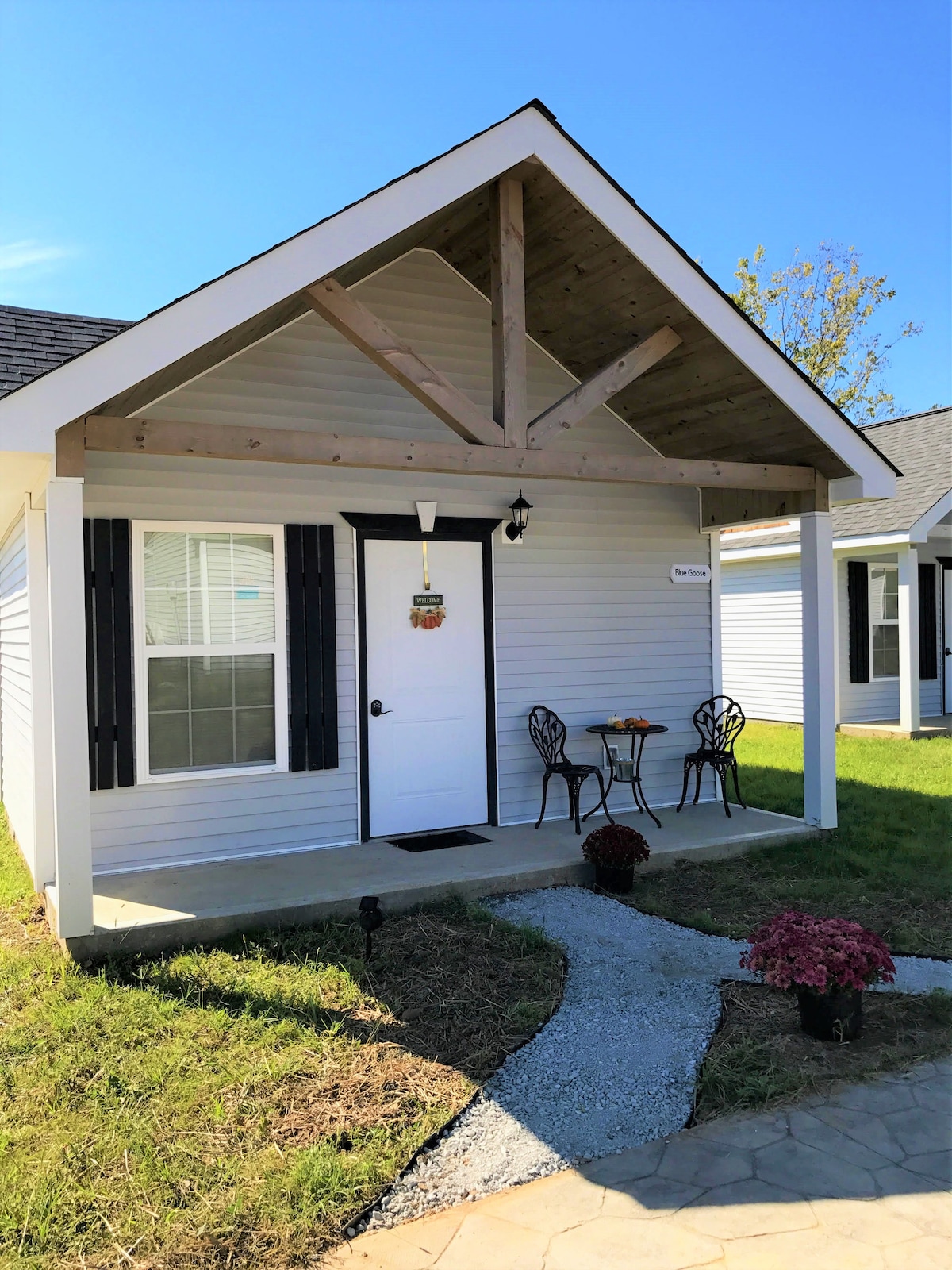 A charming cottage exterior is showcased with a covered porch supported by a wooden beam. Two elegant chairs are positioned at a small table, inviting relaxation. A walkway of gravel leads to the door, surrounded by well-maintained grass and flowering plants.