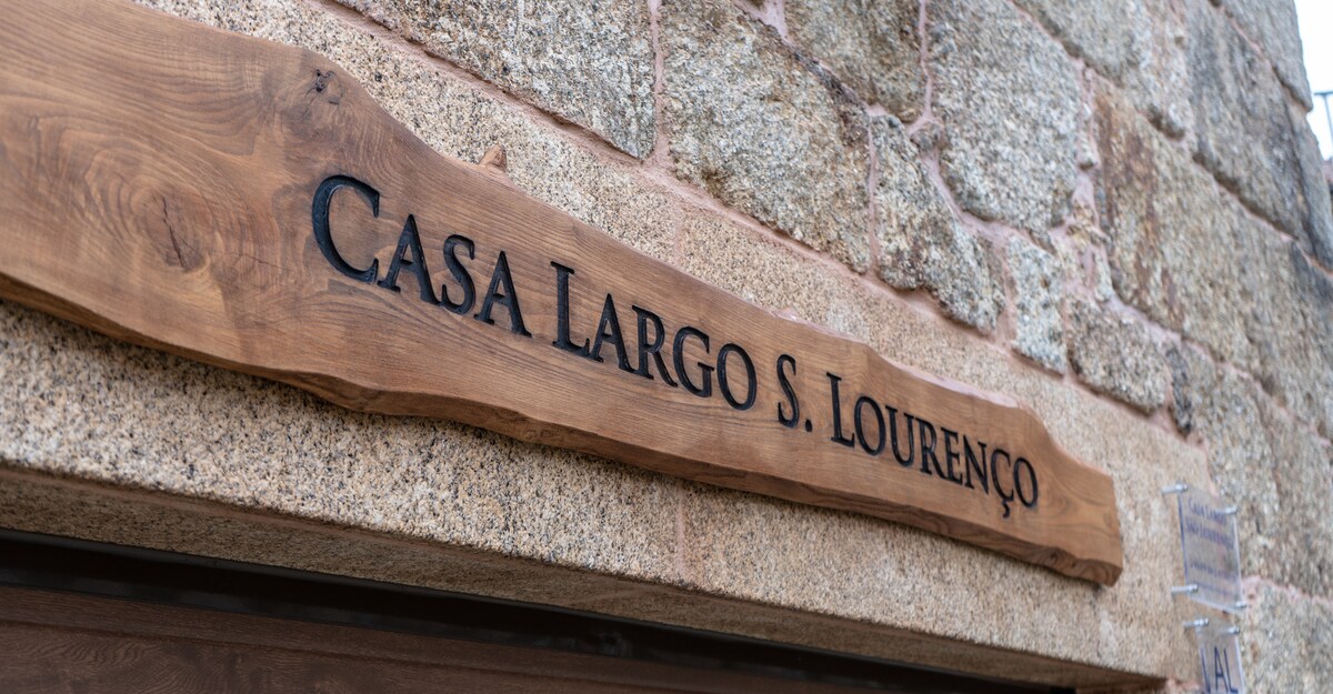 A rustic wooden sign is prominently displayed, featuring the text 'Casa Largo S. Lourenço' carved into the surface. The backdrop consists of weathered stone, adding a touch of character to the entrance.
