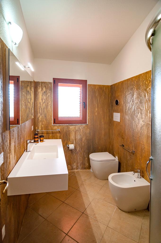 The bathroom is designed with a modern aesthetic, featuring a wall-mounted sink and a toilet alongside a bidet. Natural light enters through two windows, and textured walls create a unique backdrop. A glass shower enclosure stands in the corner, offering a clean and functional space.