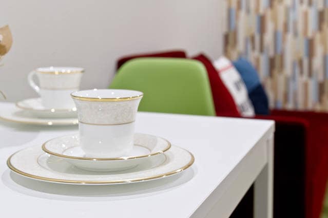 Delicate porcelain dinnerware is arranged on a table, featuring a cup, saucer, and plate with subtle gold accents. Bright seating in the background adds a pop of color, complementing the clean, modern aesthetic of the space.