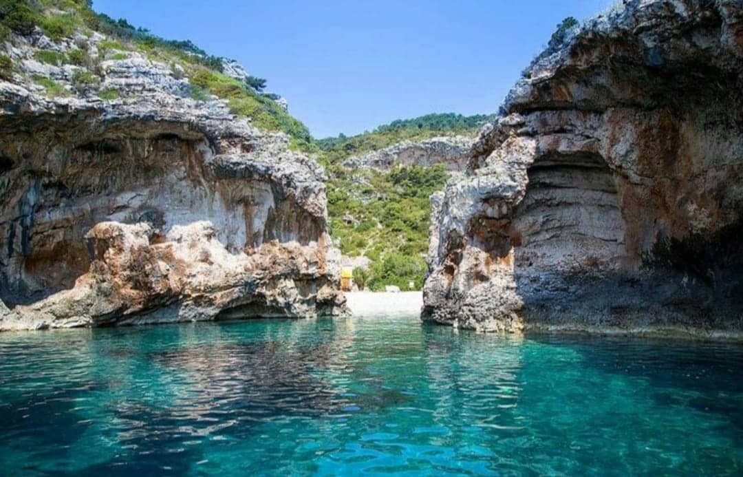 The image showcases a secluded cove surrounded by rugged rock formations, with clear turquoise waters gently reflecting the sunlight. Lush greenery is visible on the hillsides, providing a natural backdrop. A sandy beach is faintly visible in the distance, inviting for a tranquil escape.