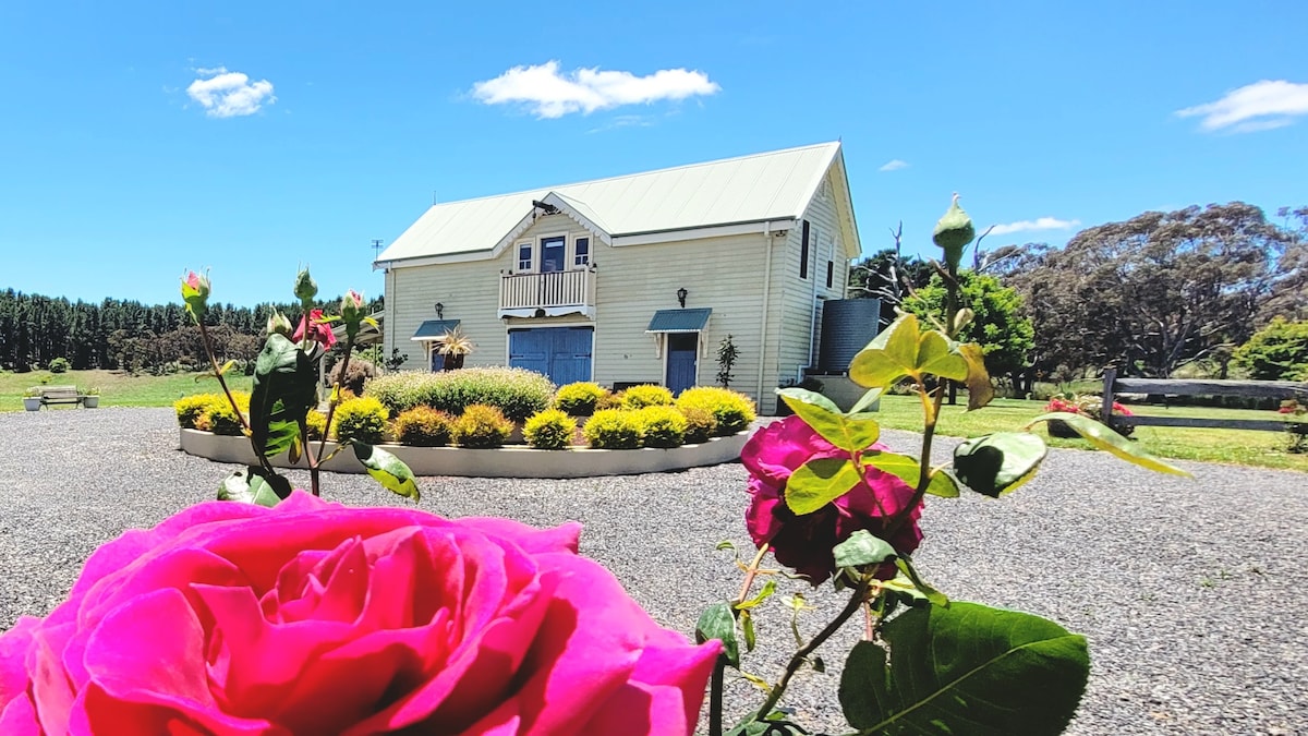 The cottage is framed by blooming roses in vibrant pink and soft yellow. A spacious building with a light-colored façade and a metal roof features a blue door and shutters. Lush landscaped gardens with seasonal flowers are arranged around a gravel path, complemented by a clear blue sky.