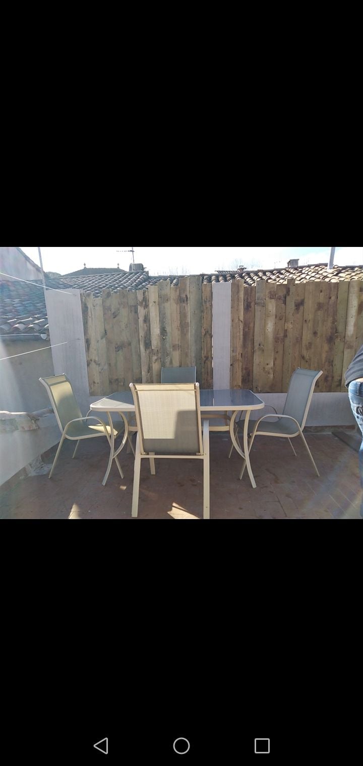 An outdoor seating area is presented, featuring a round table surrounded by four chairs. Sunlight casts reflections on the furniture, enhancing the ambience of the wooden fence in the background, which provides privacy and a natural setting.