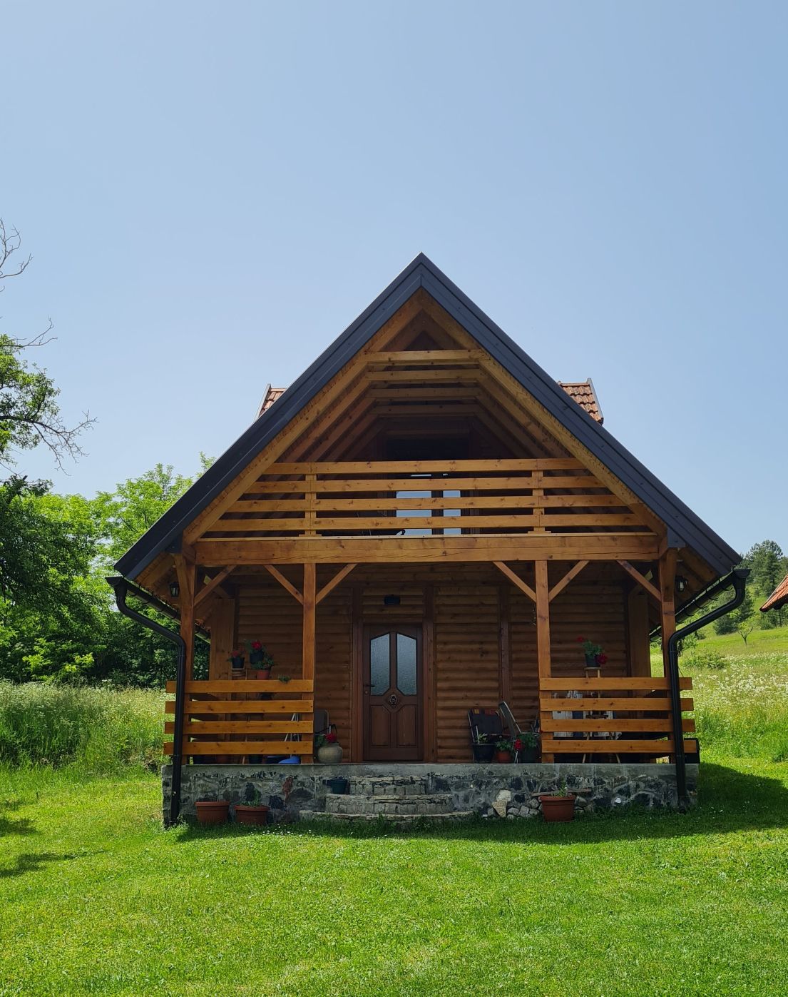 A wooden house displays a triangular roof and balconies adorned with decorative planters. The entrance is framed by a stone foundation, surrounded by green grass and trees, enhancing the natural setting of the home.