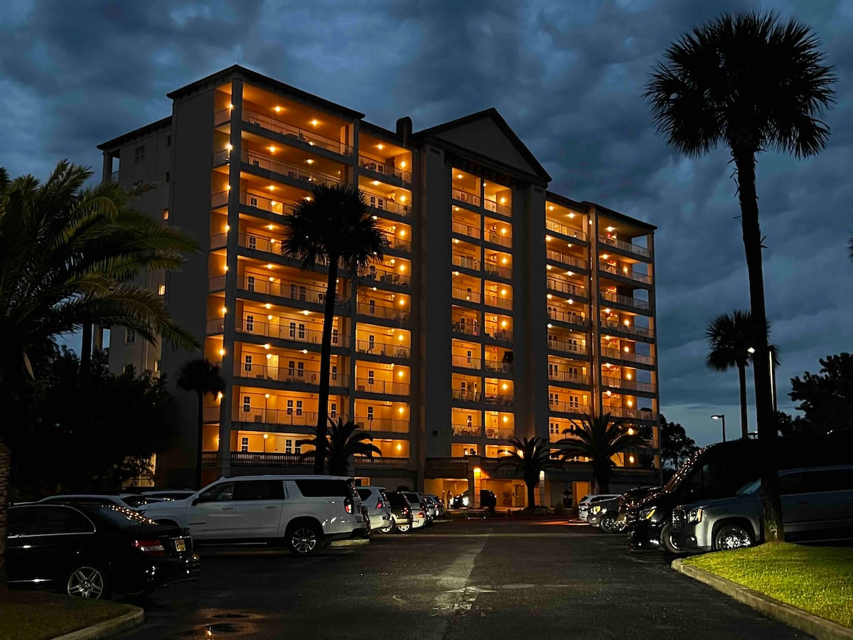 A well-lit corner unit condominium is showcased at dusk, displaying multiple balconies with warm glow lights. Palm trees line the entrance, and a parking area is visible in the foreground, featuring parked vehicles. The building's architecture is highlighted against a cloudy evening sky.