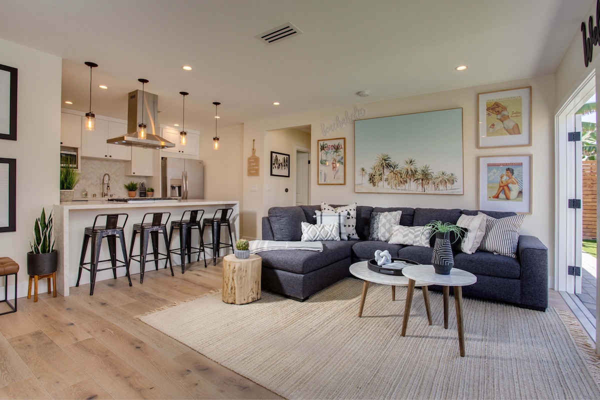An open living area features a modern sectional sofa and wooden accent tables. Bar stools line the kitchen counter, which overlooks the space. Artwork adorns the walls, complemented by neutral tones and natural light from the adjacent outdoor area. A woven rug adds warmth to the hardwood floor.