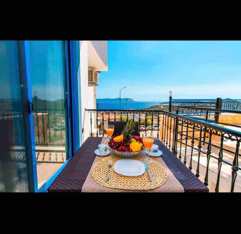 A balcony table is set with a fruit arrangement, two glasses of orange juice, and a decorative placemat. Expansive views of the sea and surrounding landscape are visible through large glass doors, providing a bright and inviting atmosphere for outdoor dining.