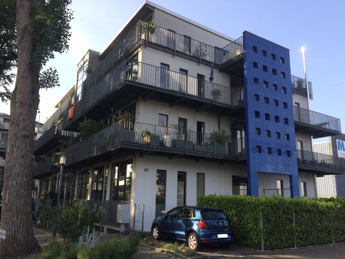 A modern, multi-story building is showcased with multiple balconies, allowing for outdoor spaces. A blue accent wall adds a distinctive feature. A car is parked in front, while greenery lines the base of the structure, enhancing the overall appearance.