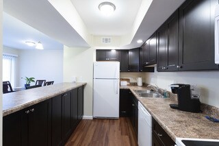 A modern kitchen is equipped with dark cabinetry and a granite counter. The space contains a refrigerator, sink, and coffee maker. Soft lighting enhances the airy feel, while a dining area is visible in the background, furnished with a wooden table and chairs.