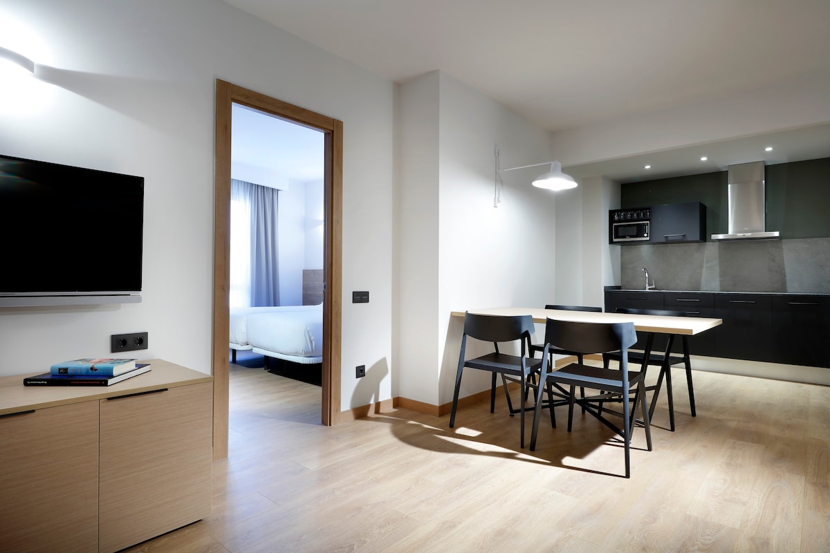 A modern and open living space includes a dining area with a wooden table and four black chairs. A mounted TV is positioned on a light-toned cabinet, while a sleek kitchen with dark cabinetry and a hood is visible in the background. Soft lighting enhances the minimalist design.