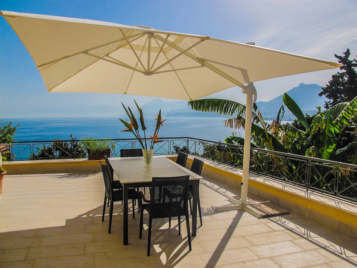 An outdoor dining area is featured under a large umbrella, designed for shade. A spacious table is surrounded by six black chairs, offering a panoramic view of the sea and surrounding greenery, enhancing the outdoor experience.