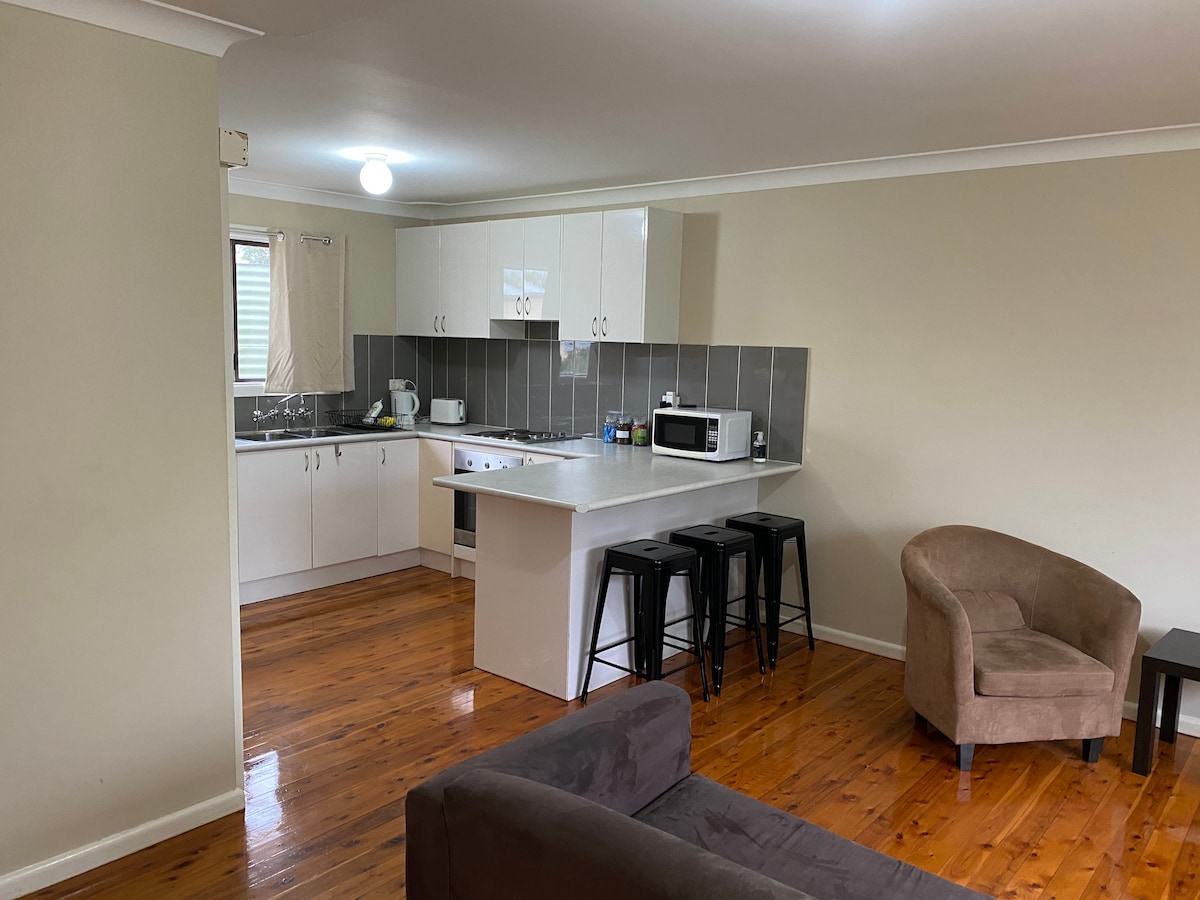 An open kitchen and living area features modern cabinetry and stainless-steel appliances. A small dining area is highlighted by high-backed stools. Wooden flooring provides a warm tone, complemented by a brown armchair and a soft gray sofa, creating a comfortable space for relaxation.