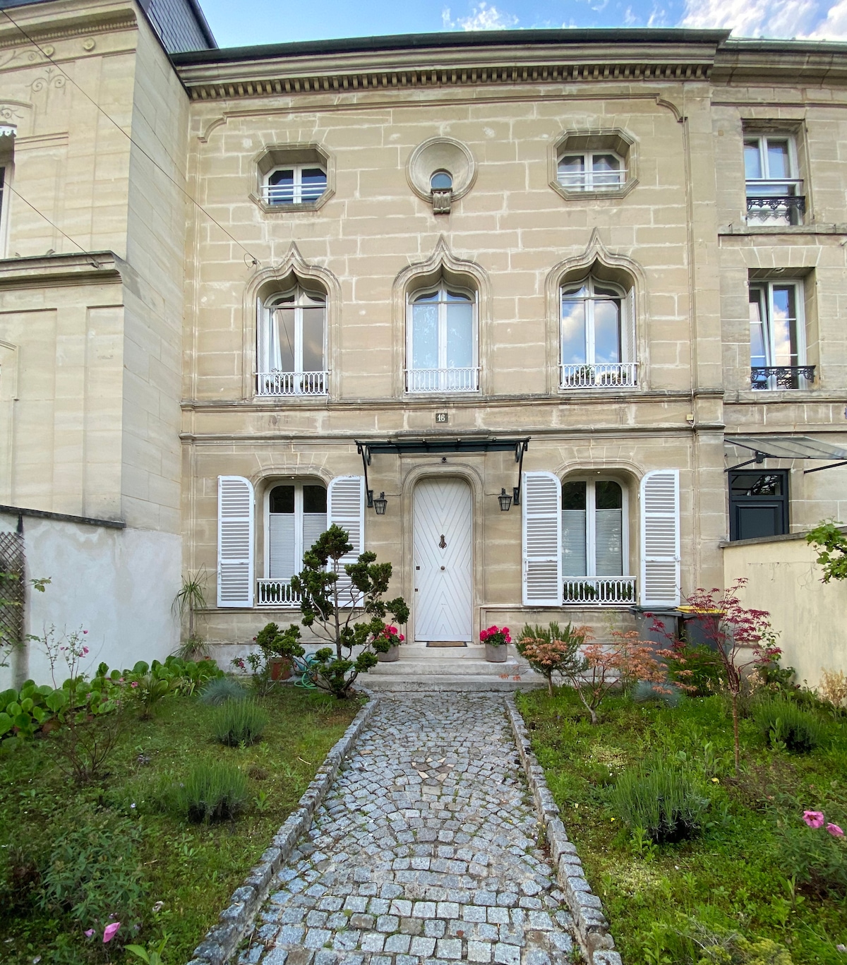 A charming facade of a historical building is presented, featuring ornate windows with decorative arches. A welcoming entryway is accentuated by a simple door framed by two lanterns. A cobblestone path leads through the garden, filled with lush greenery and vibrant flowers.