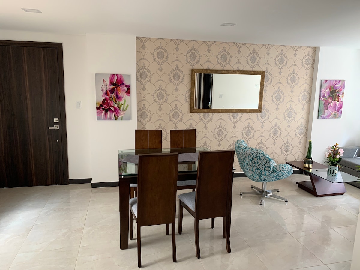 The living area features a modern design with a mirrored wall, a glass dining table surrounded by four wooden chairs, and an upholstered accent chair. Floral artwork adorns the walls, adding a touch of color to the neutral tones of the space.