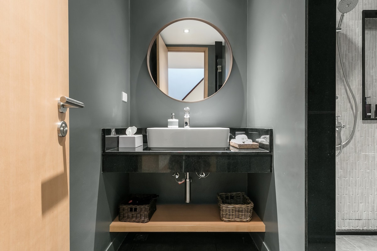 A modern bathroom is presented with a large oval mirror above a sleek black countertop. A spacious white basin is centered on the counter, complemented by neatly arranged toiletries. Below, wooden shelves hold woven baskets, creating a clean and organized space.