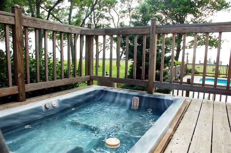 A hot tub is situated on a wooden deck, surrounded by a wooden railing for privacy. The water is bubbling gently. In the background, an expansive lawn and trees are visible, creating a serene outdoor environment.