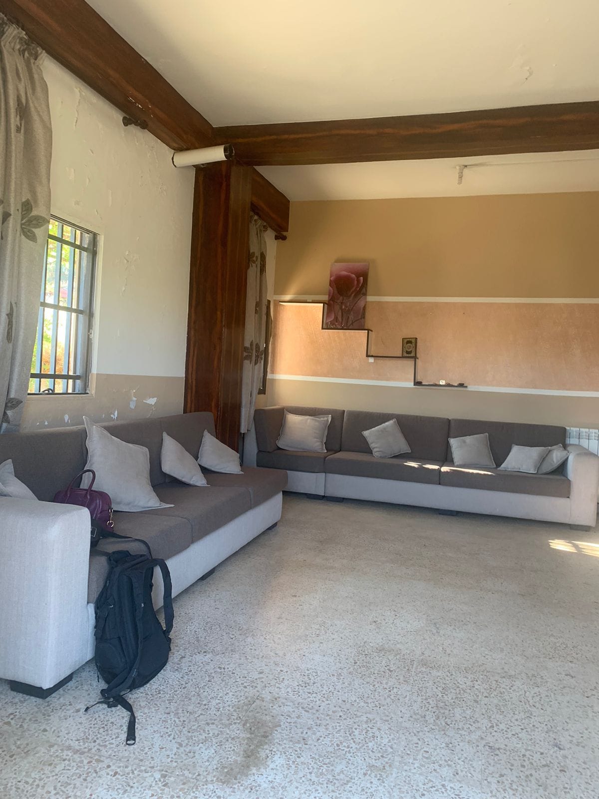 A spacious living room features two large sofas with decorative cushions arranged around a central space. Natural light floods in through a window, illuminating the textural walls. An artwork piece is displayed on the wall, adding a subtle touch of character.