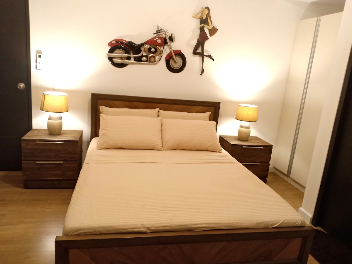 A cozy bedroom features a queen size bed with light-colored linens, flanked by two bedside tables with lamps. A decorative motorcycle wall art piece adds character, alongside a stylish figure. A mirrored wardrobe stands against the far wall, enhancing the room's spacious feel.
