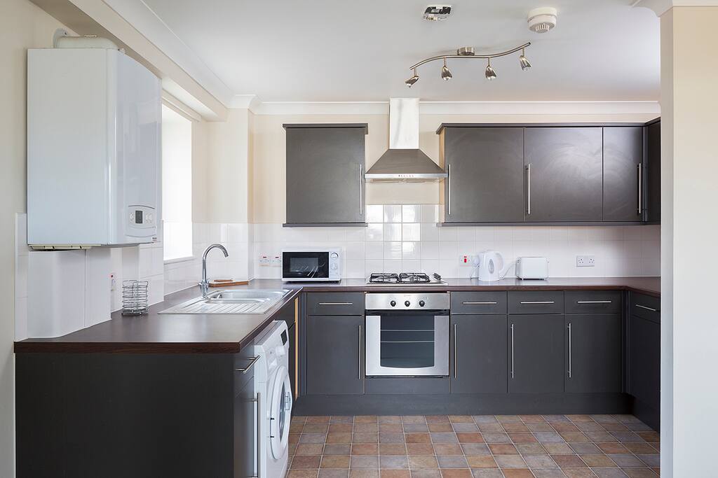 Modern kitchen with fridge, cooker, washing machine, microwave, kettle and toaster, plates, pots and pans, glasses, mugs and cutlery