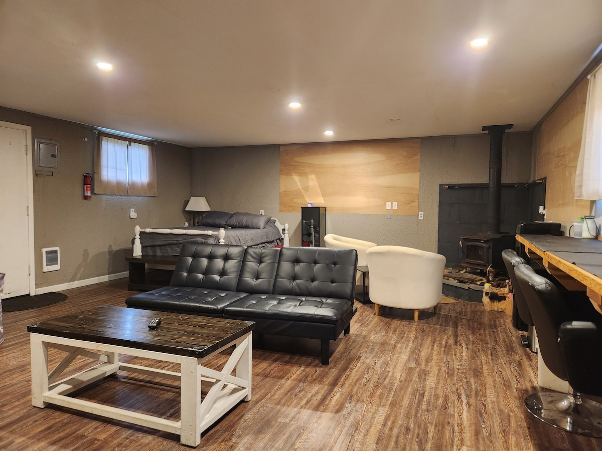 The studio apartment interior is spacious and features a black couch and a white armchair, arranged around a white coffee table. A full-size bed with gray bedding is positioned against a wood-paneled wall. The area is illuminated by overhead lighting.