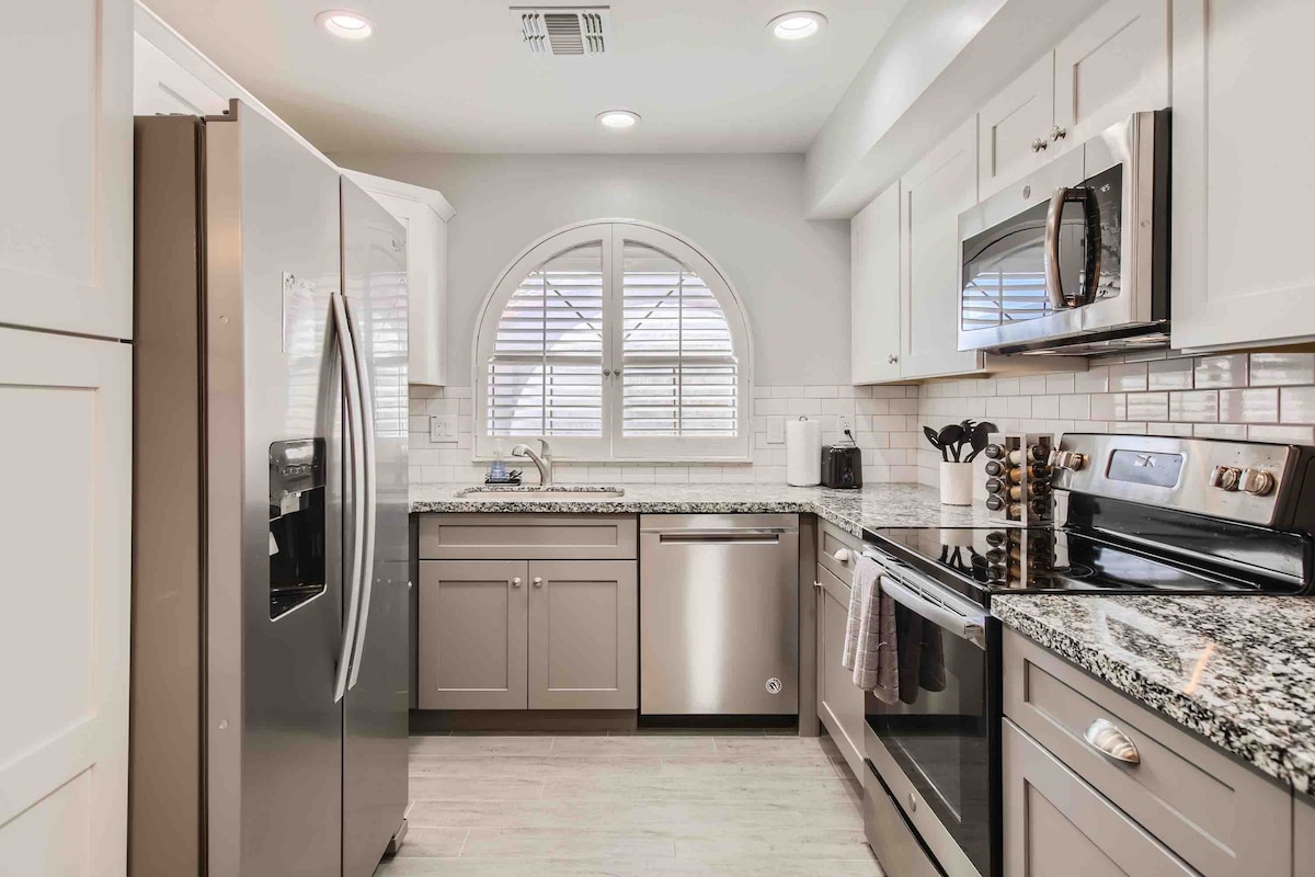 The kitchen features modern stainless steel appliances, including a refrigerator and oven. A granite countertop extends along the space, complemented by white cabinets. Natural light enters through an arched window, which is adorned with blinds, creating a functional and inviting cooking environment.