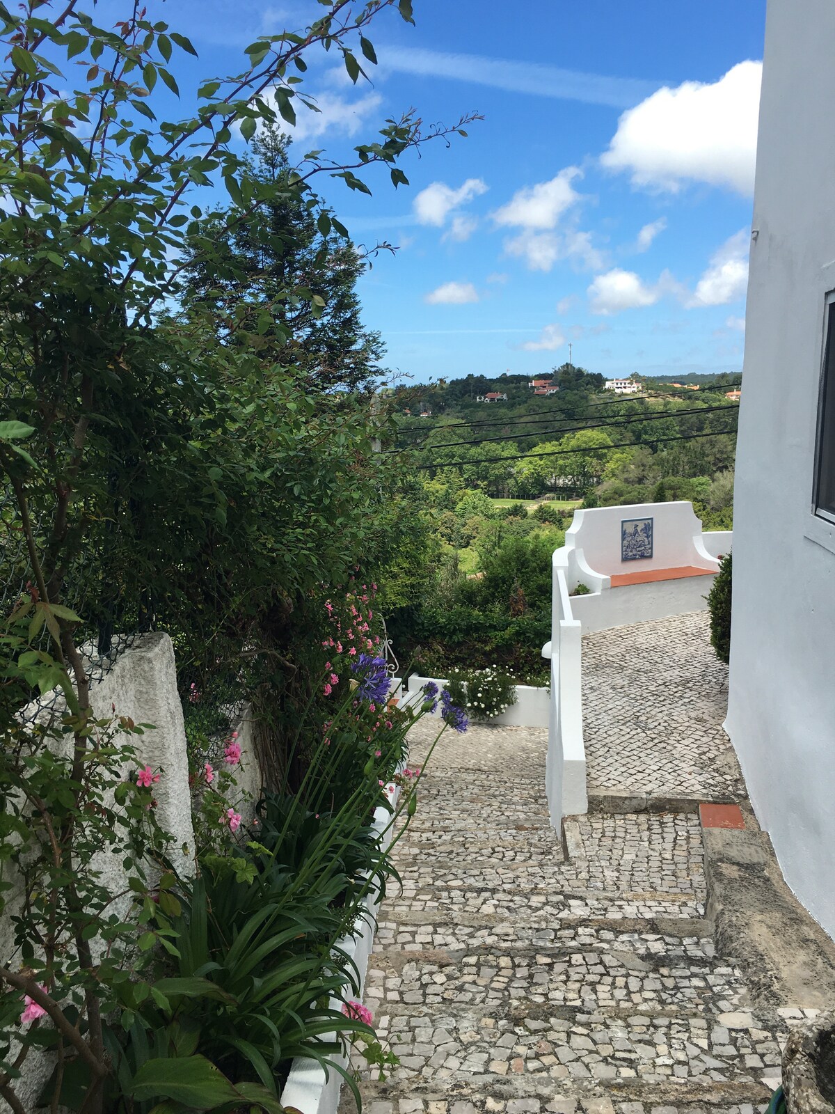 A winding stone staircase is flanked by lush greenery and colorful flowers, leading down towards a picturesque view of the countryside. The clear blue sky creates a bright atmosphere, enhancing the serene landscape that unfolds below.