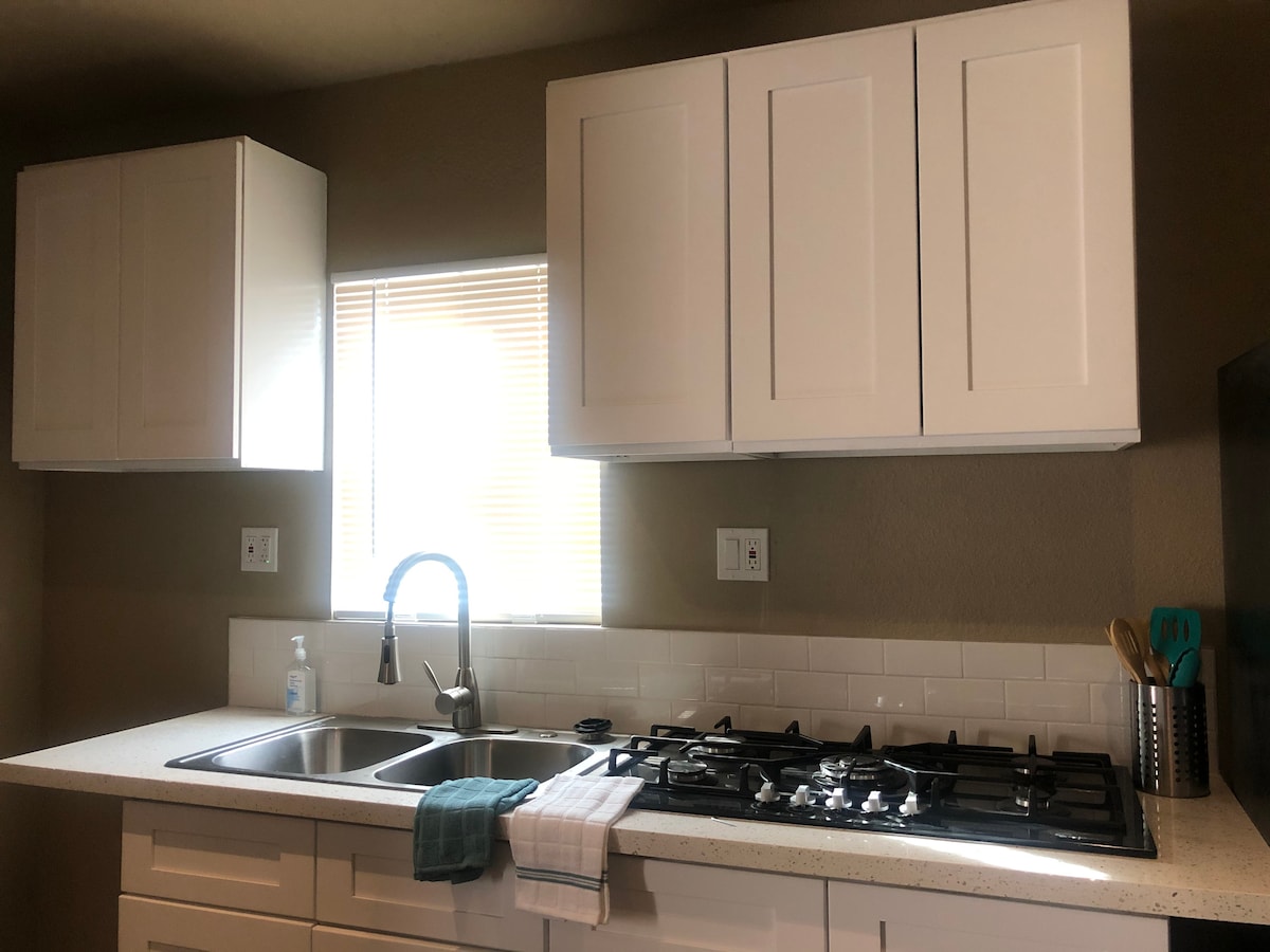 A modern kitchen is equipped with a double sink and a gas stove, featuring white cabinetry that contrasts with the neutral wall color. Natural light enters through a window, illuminating a clean countertop, where utensils and a dish towel are neatly placed.