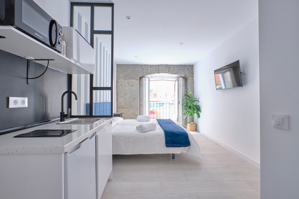 A modern studio features a spacious layout with a double bed dressed in white linens and towels arranged neatly. A kitchenette area with a microwave and sink is included, alongside a flat-screen TV mounted on the wall. Natural light floods through the window, enhancing the relaxed atmosphere.