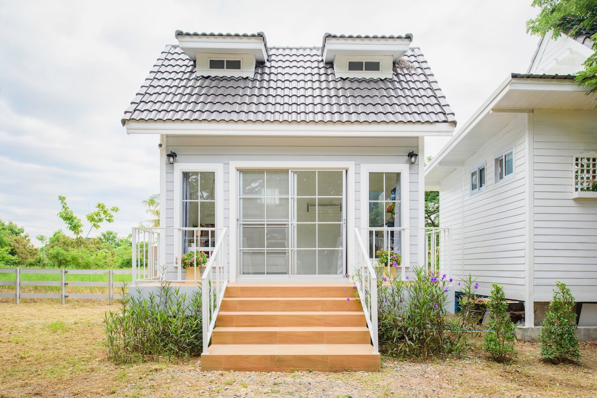 A charming white cottage features a symmetrical roof and large glass doors that open onto a wooden staircase. Lush greenery and flower beds adorn the exterior, creating an inviting entrance. The surrounding landscape includes open fields and a cloudy sky.