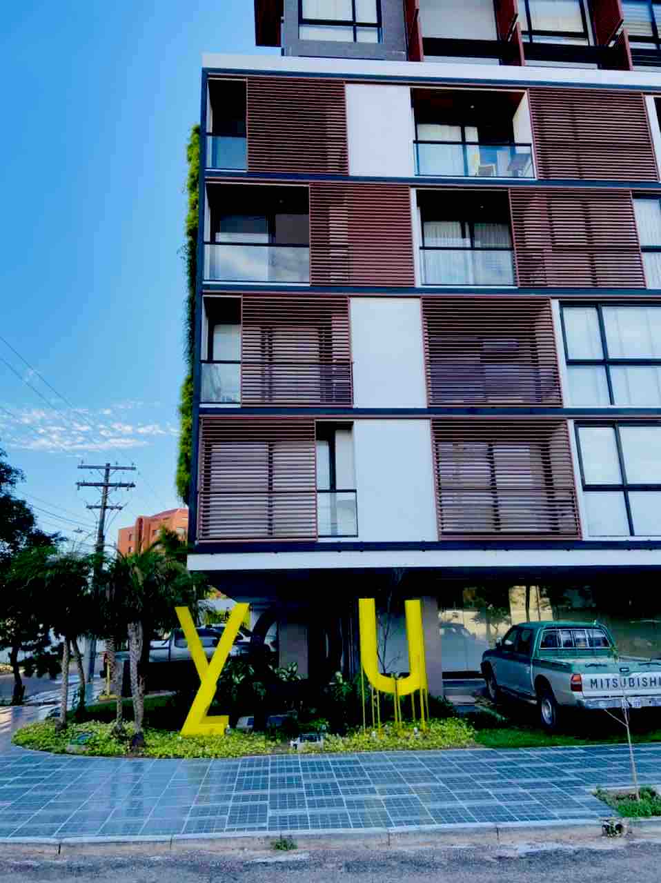 The building's exterior features modern architectural lines, highlighted by large windows and contrasting materials. Greenery surrounds the base, while a brightly colored sign stands out in the foreground. A parked vehicle is visible, contributing to the contemporary urban setting.
