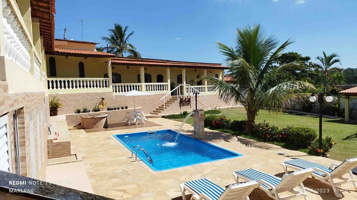 A clear view of a serene pool surrounded by sun loungers is presented. Lush greenery and palm trees frame the space, while a two-story building with a terracotta roof is visible in the background, providing an inviting atmosphere.