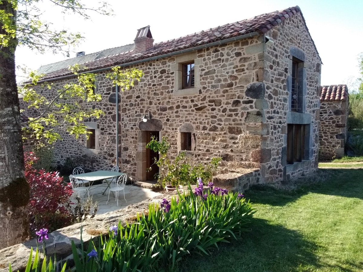 The rustic stone exterior of a renovated farmhouse is framed by green grass and colorful flowers. A small terrace with a white table and chairs is visible, inviting outdoor relaxation. Large windows and a traditional roof add to the charm of this countryside retreat.
