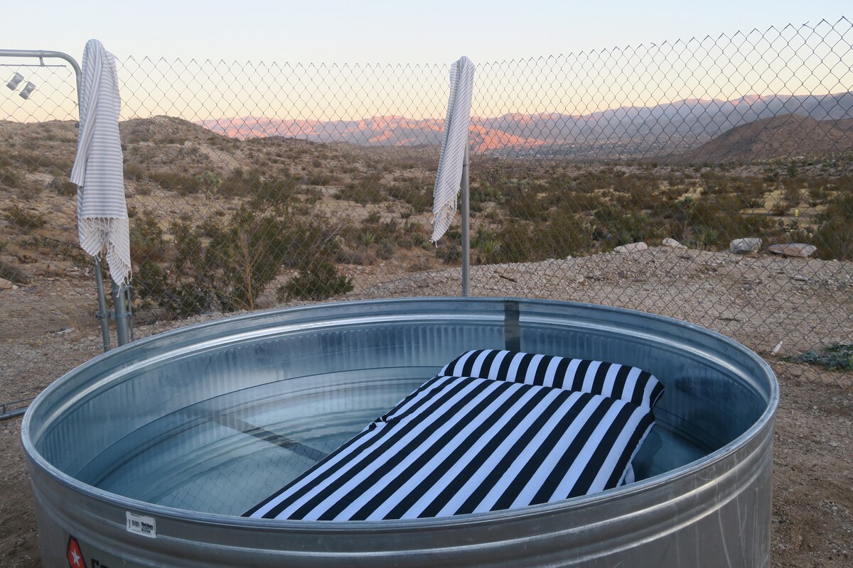 A cowboy tub filled with water features a black and white striped cushion. Surrounded by a chain-link fence, the tub offers expansive views of the high desert landscape and distant mountains, with a golden hue reflecting from the sunset.